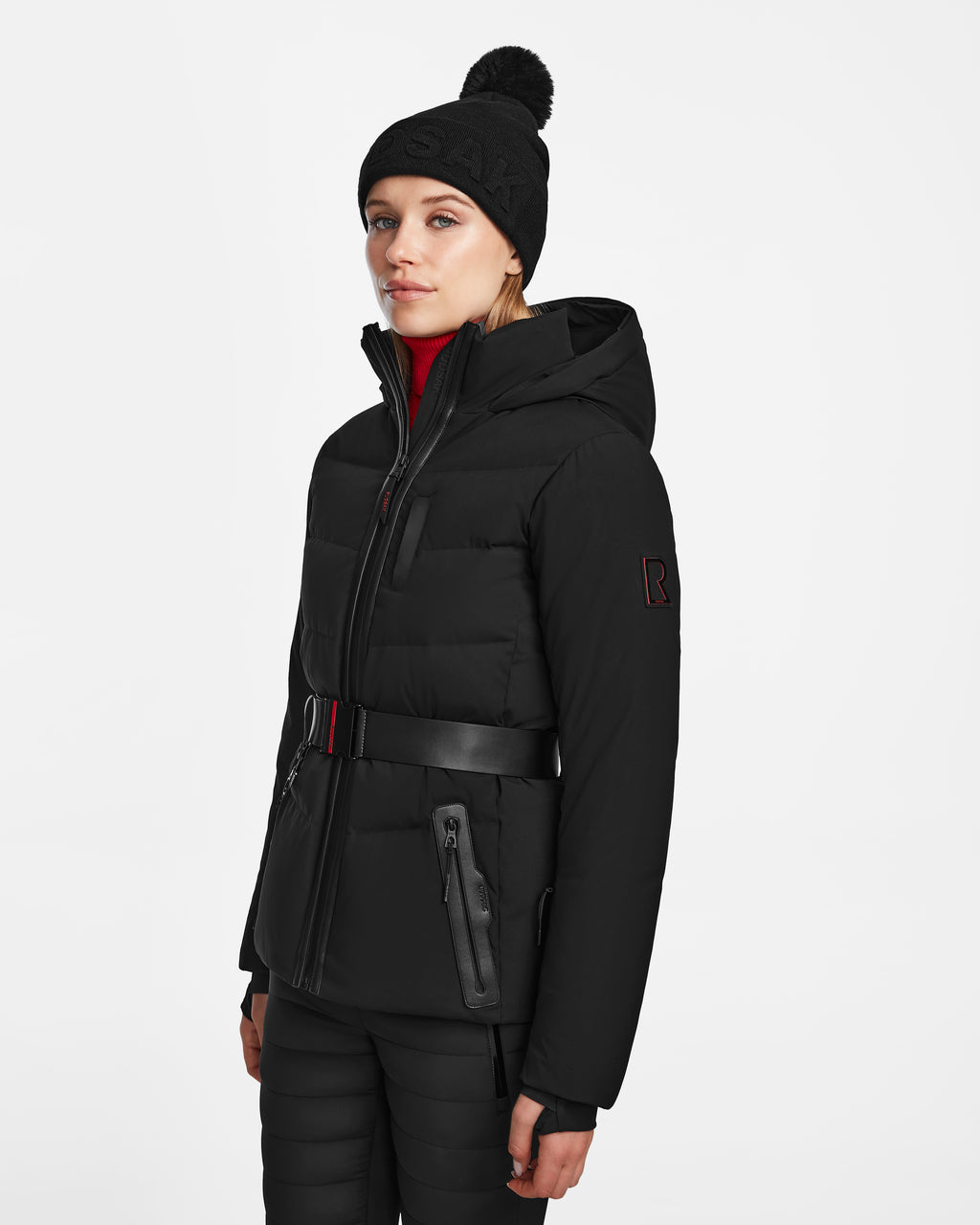 A woman wears the Rudsak ALAIA - 8124816 black quilted ski jacket with a belt, paired with black pants and a pom-pom beanie, standing against a plain white background.
