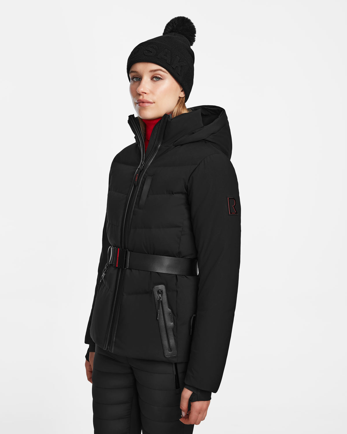 A woman wears the Rudsak ALAIA - 8124816 black quilted ski jacket with a belt, paired with black pants and a pom-pom beanie, standing against a plain white background.