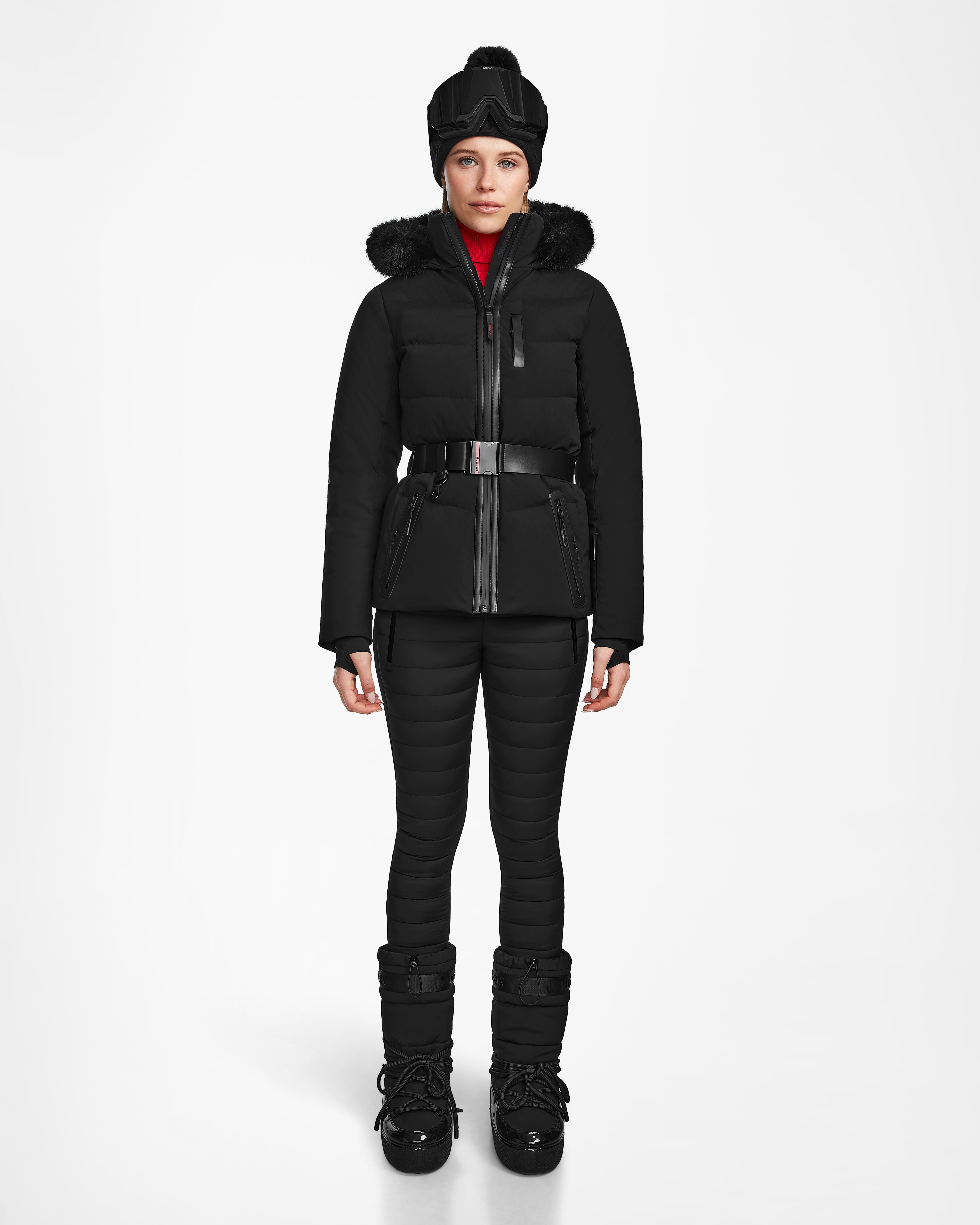 A person stands facing forward, wearing the Rudsak ALAIA - 8124816 Black women’s quilted ski jacket with a fur-trimmed hood, matching pants, winter boots, wide belt, black hat, and RECCO rescue technology for cold weather safety.