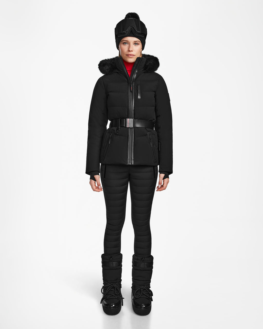 A person stands facing forward, wearing the Rudsak ALAIA - 8124816 Black women’s quilted ski jacket with a fur-trimmed hood, matching pants, winter boots, wide belt, black hat, and RECCO rescue technology for cold weather safety.