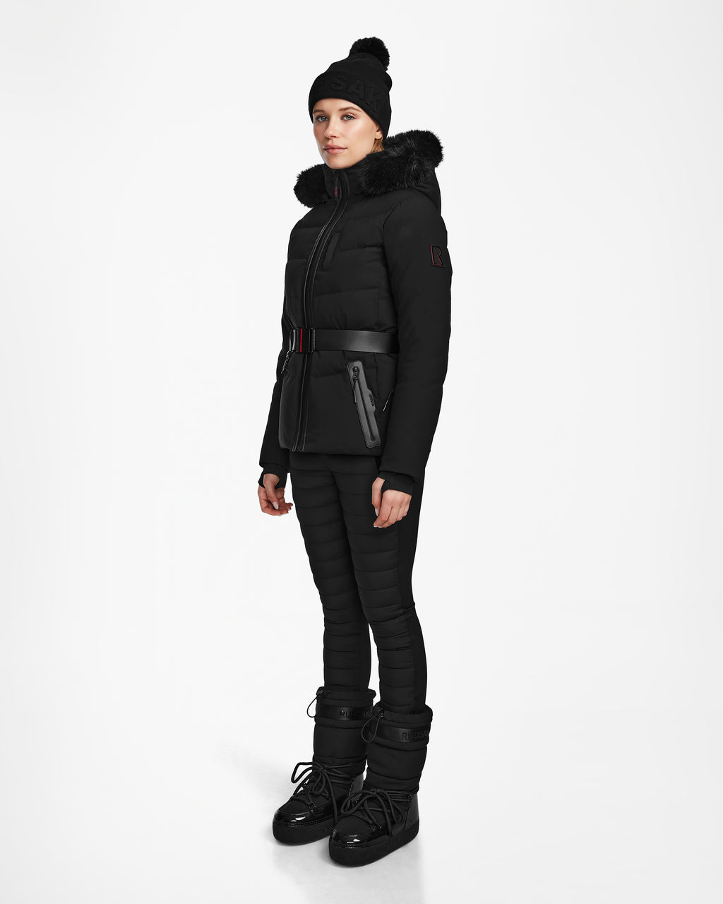 A woman wears the Rudsak ALAIA - 8124816 Black womens quilted ski jacket with a fur-trimmed hood, belted puffer design, matching pants, black beanie, and winter boots—ideal for the slopes—against a white background.