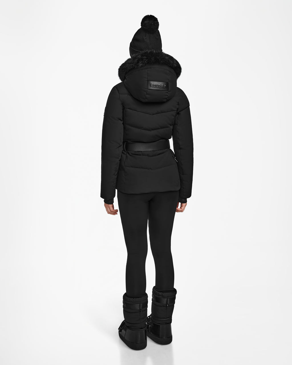 A person faces away against a white background, wearing the Rudsak ALAIA - 8124816 Black womens quilted ski jacket with fur-trimmed hood, paired with black leggings, boots, and a black knit hat with a pom-pom.