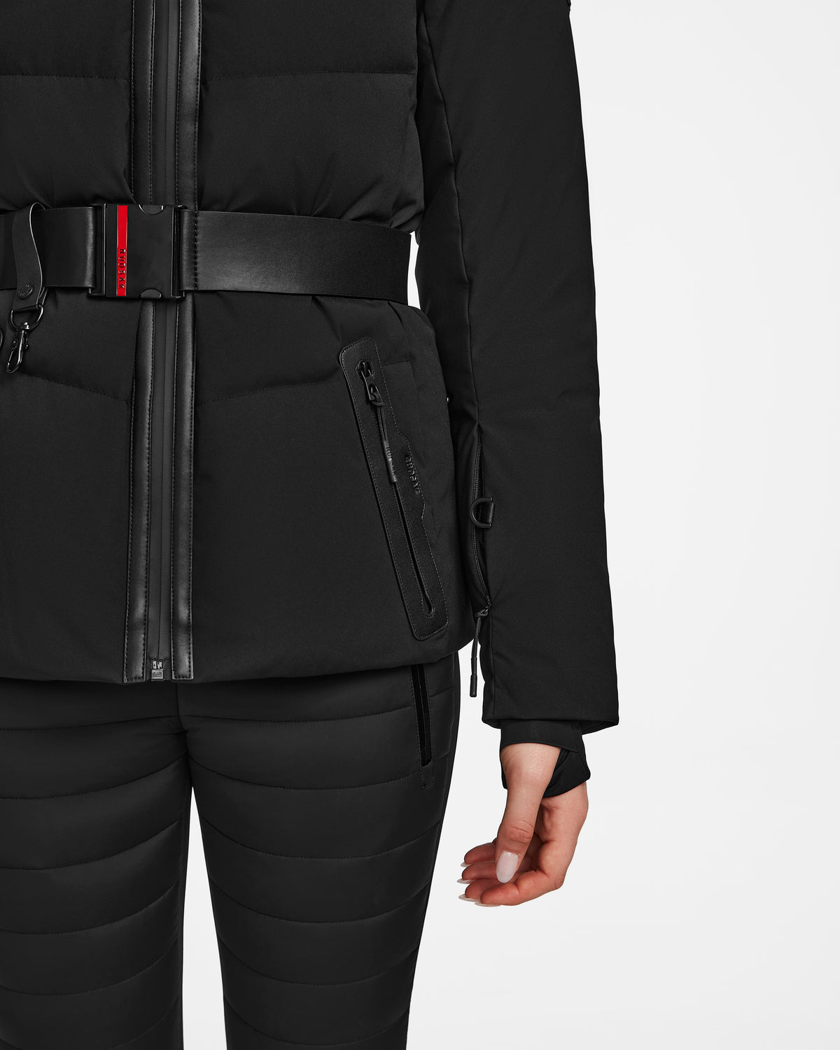A person models the Rudsak ALAIA - 8124816 Black womens quilted ski jacket with a zipper and black belt, paired with matching black pants, shown from the waist down against a white background.