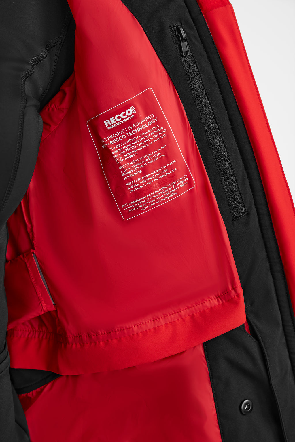 Close-up of the inside of the Rudsak ALAIA - 8124816 Flame Red ski jacket, featuring a label for RECCO rescue technology and a zippered chest pocket on the vibrant red lining.
