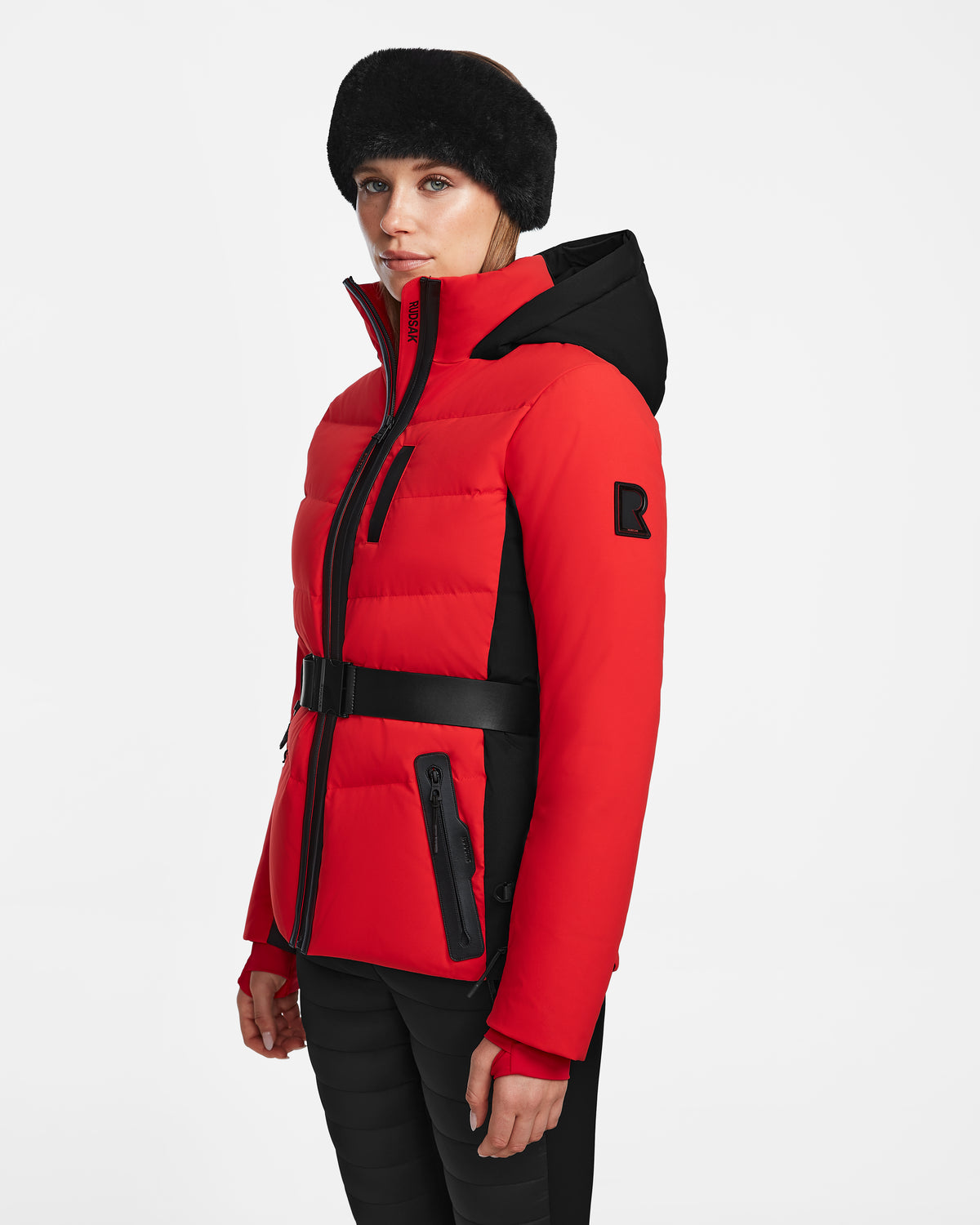 A woman wears the Rudsak ALAIA - 8124816 Flame Red puffer ski jacket with RECCO rescue tech, black pants, and a black fur hat, standing against a plain white background and looking slightly to the side.