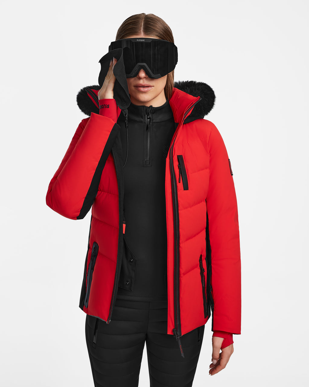 A woman wearing the Rudsak ALAIA - 8124816 Flame Red quilted ski jacket with a fur-lined hood, black ski goggles, and black pants stands against a white background, holding her goggles with one hand.