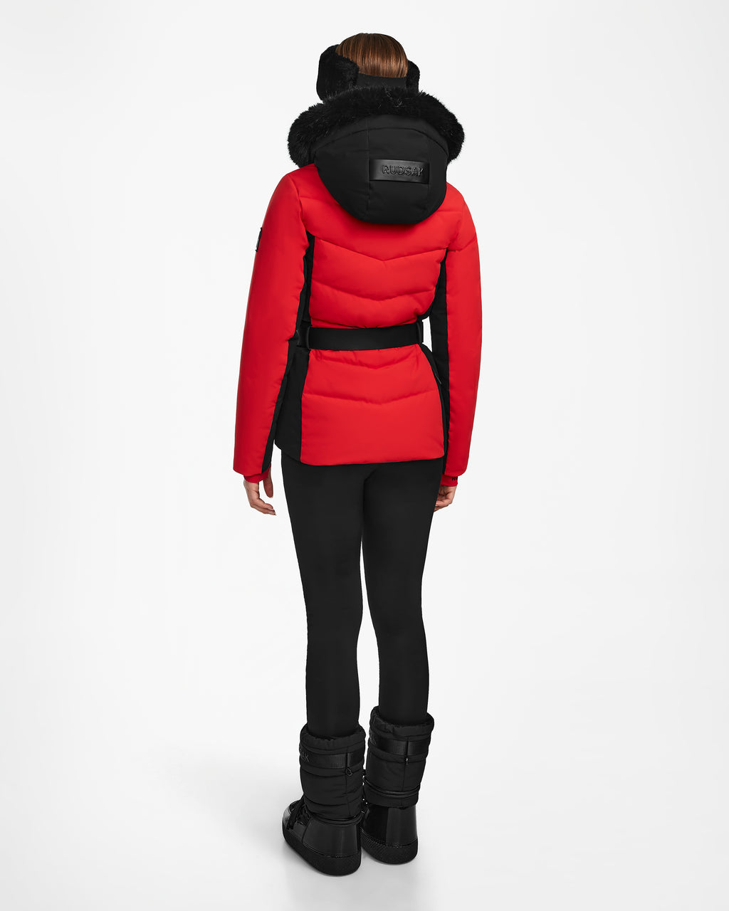 A person stands facing away, wearing the Rudsak ALAIA - 8124816 Flame Red ski jacket with a black belt, paired with black leggings and winter boots against a plain white background.