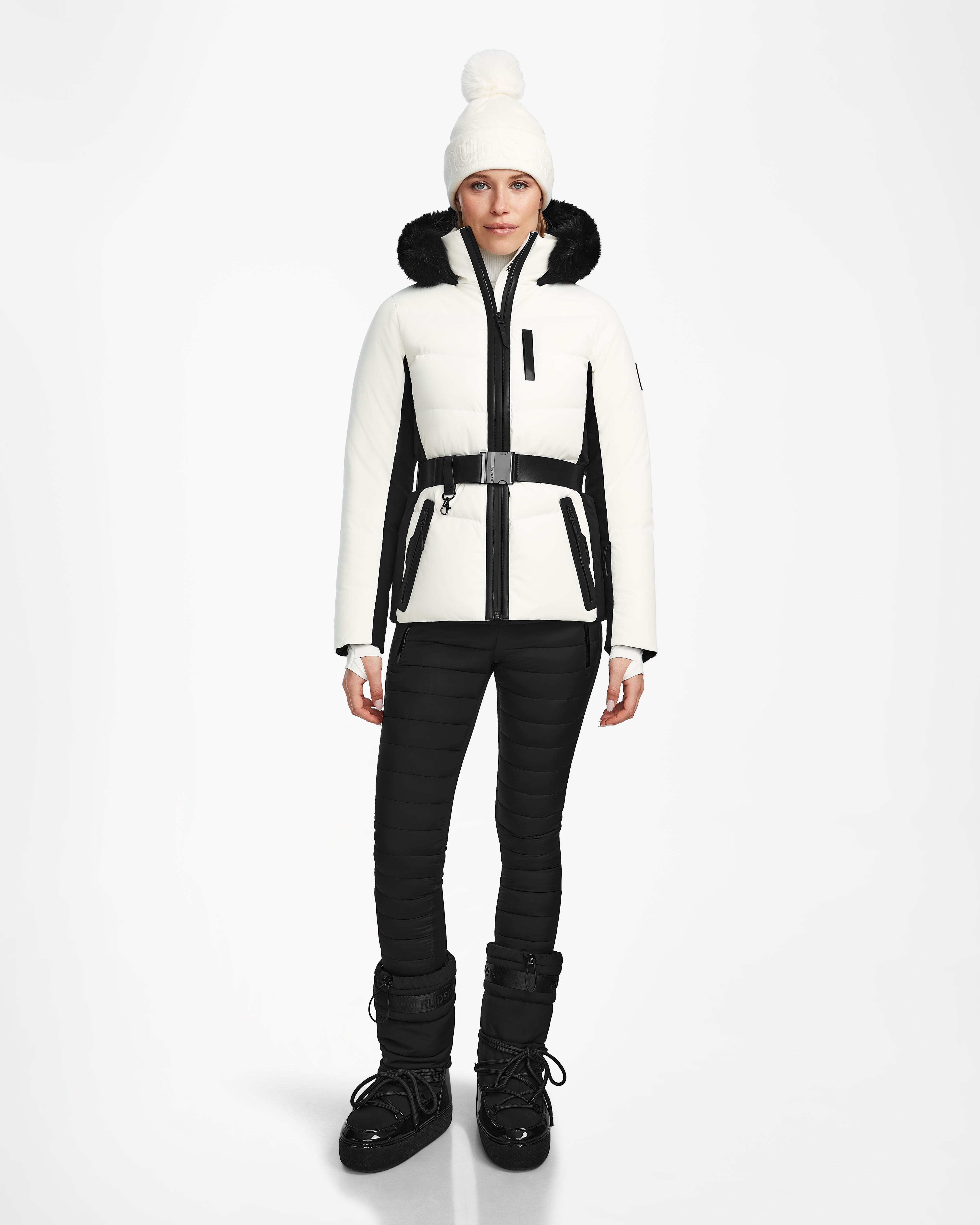 A woman faces forward against a white background, wearing the Rudsak ALAIA - 8124816 Iceland ski jacket with black accents, a white pom-pom beanie, black fitted pants, and black snow boots.