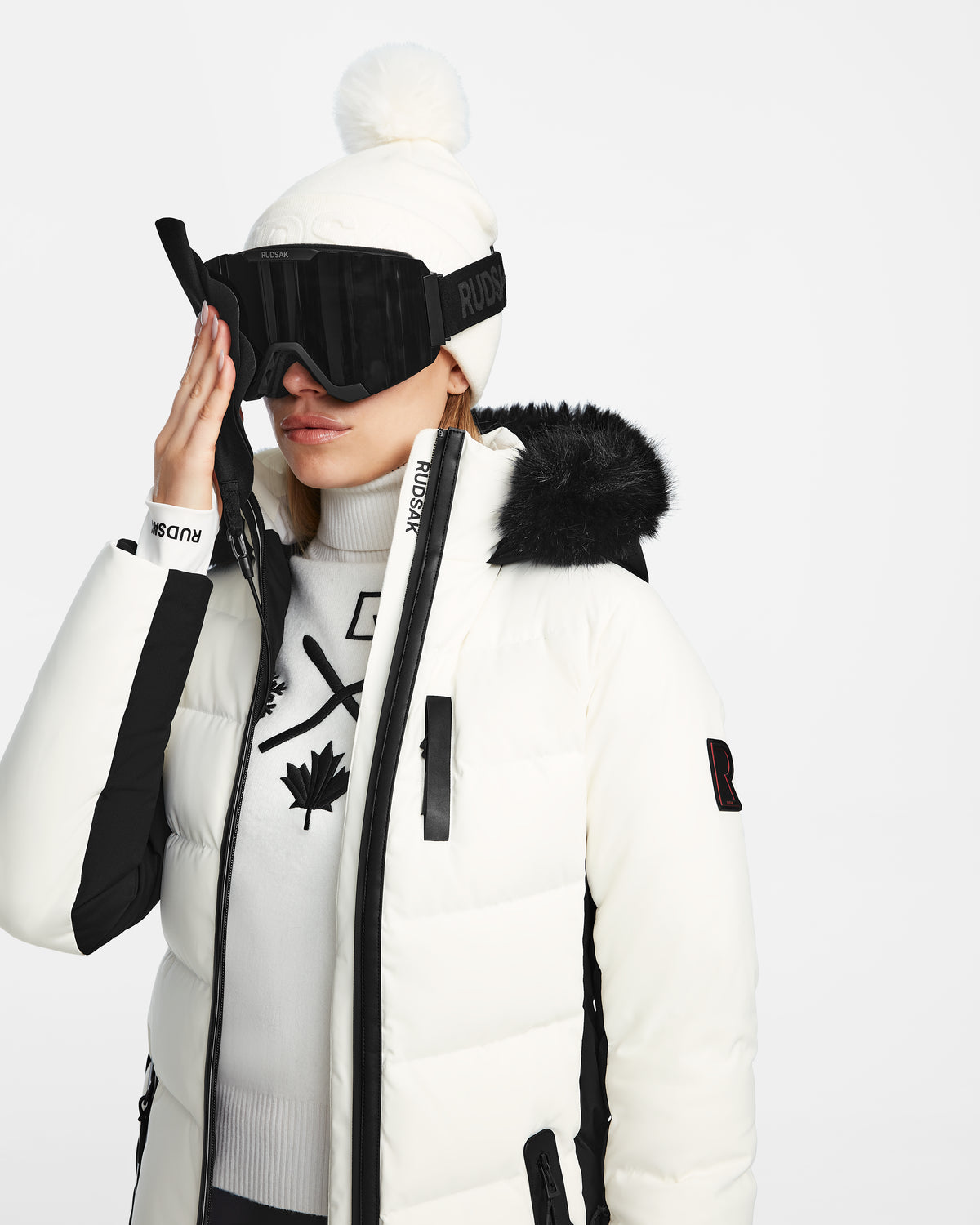 Wearing the Rudsak ALAIA - 8124816 Iceland jacket with RECCO tech, black accents, and fur-trimmed hood over a maple leaf logo sweater, plus a white pom-pom beanie and black ski goggles creates a chic winter outfit.
