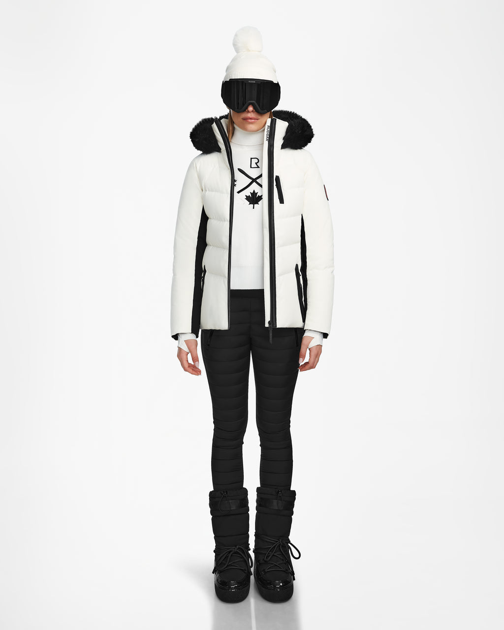 A person stands facing forward in the Rudsak ALAIA - 8124816 Iceland ski jacket with a fur-lined hood, black pants and boots, black ski gloves and goggles, plus a white beanie with pom-pom—ready to hit the slopes in style.