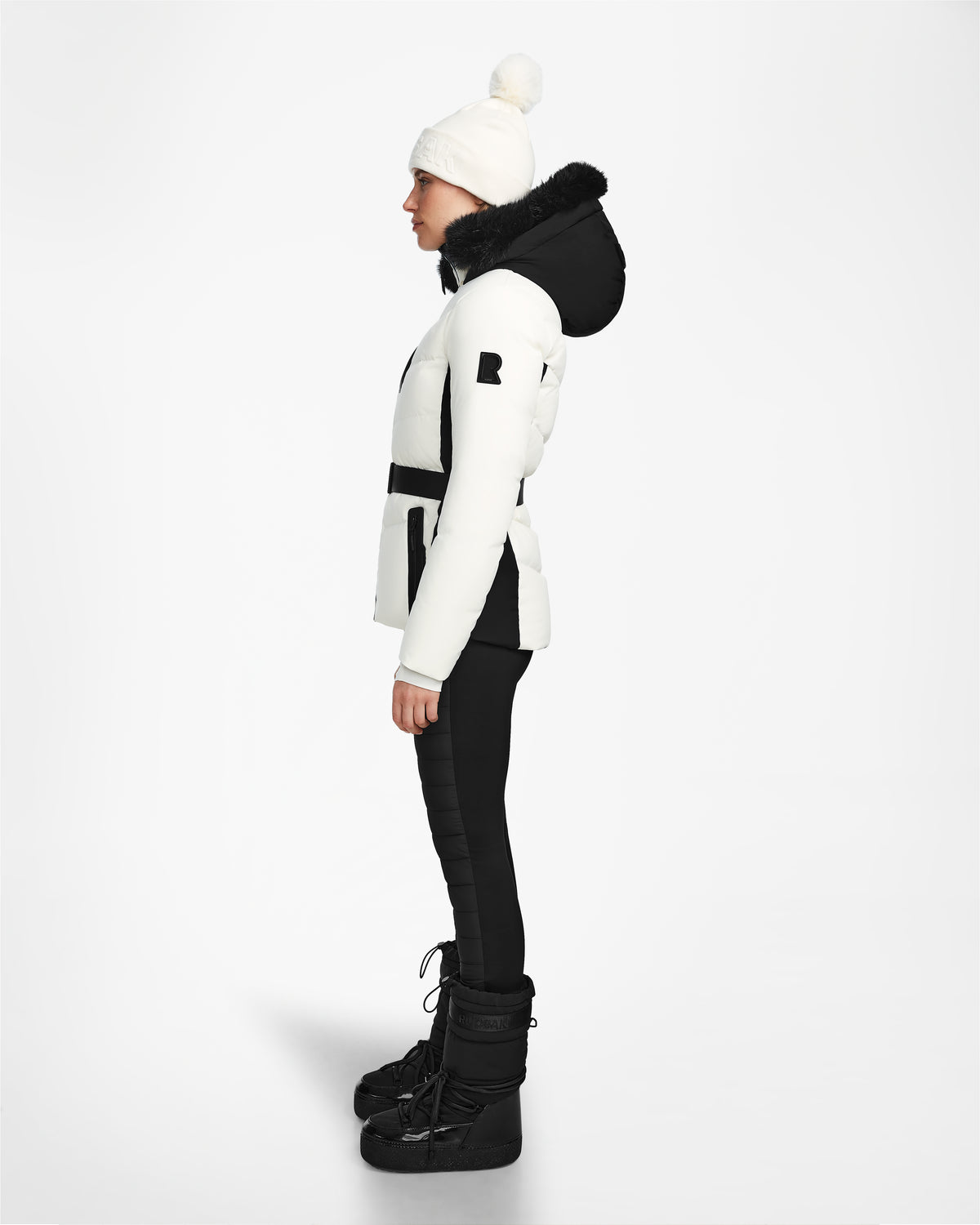 A person in profile wears the Rudsak ALAIA - 8124816 Iceland ski jacket with black accents, black pants, winter boots, and a white pom-pom beanie, standing against a plain white background.