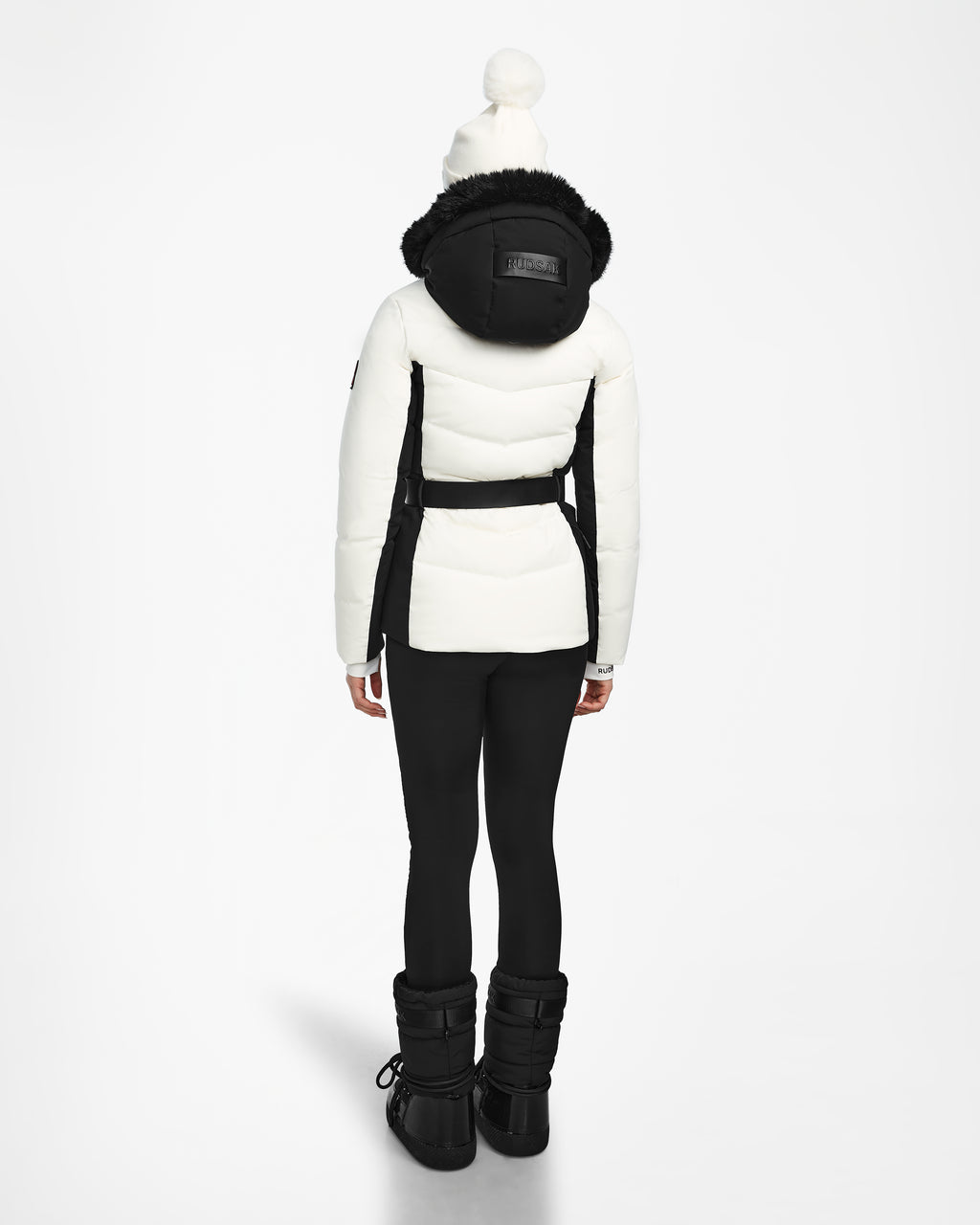 A person faces away, wearing the Rudsak ALAIA - 8124816 Iceland ski jacket with a black hood and belt, black leggings and boots, plus a white pompom beanie. The background is plain and light-colored.