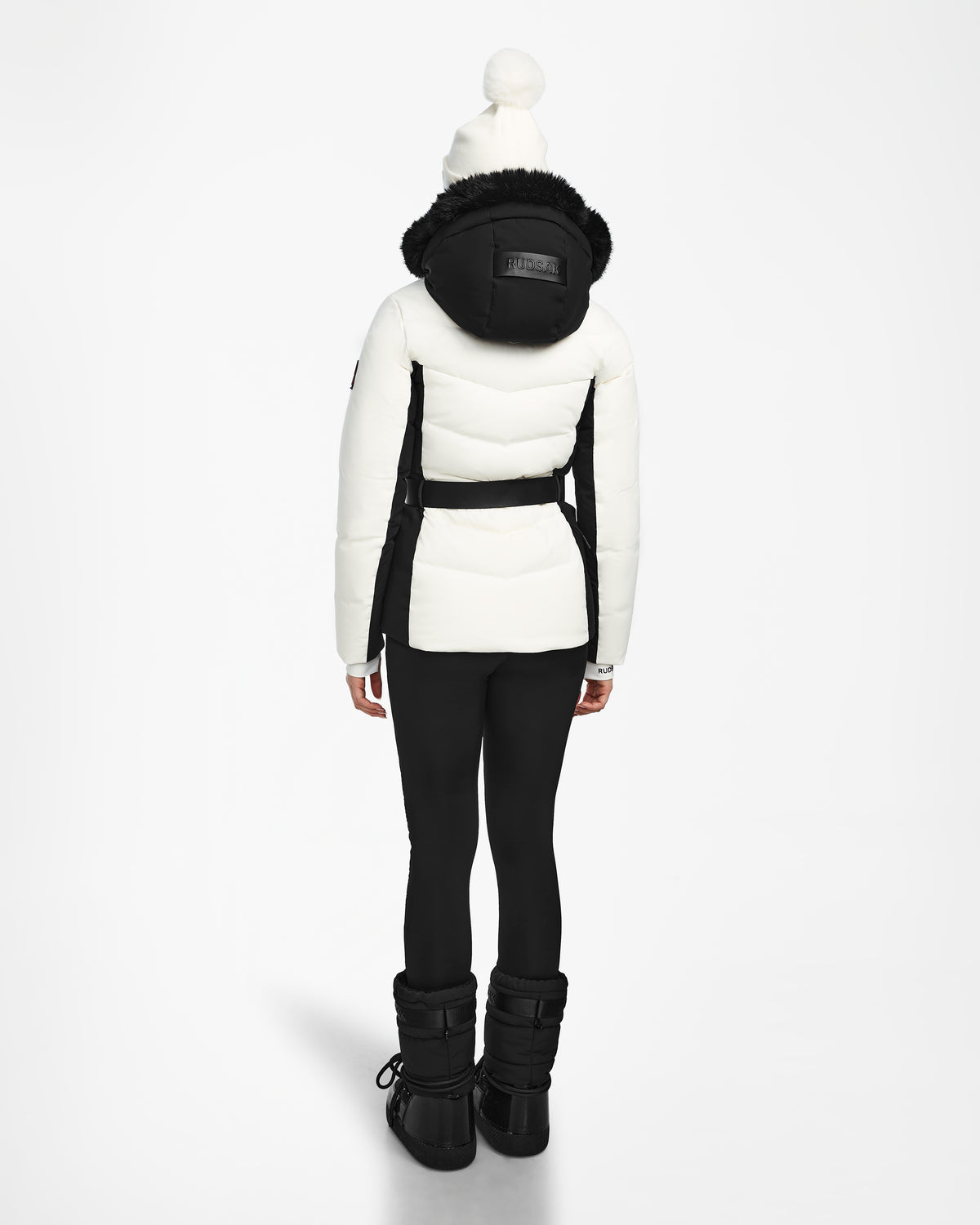 A person faces away, wearing the Rudsak ALAIA - 8124816 Iceland ski jacket with a black hood and belt, black leggings and boots, plus a white pompom beanie. The background is plain and light-colored.