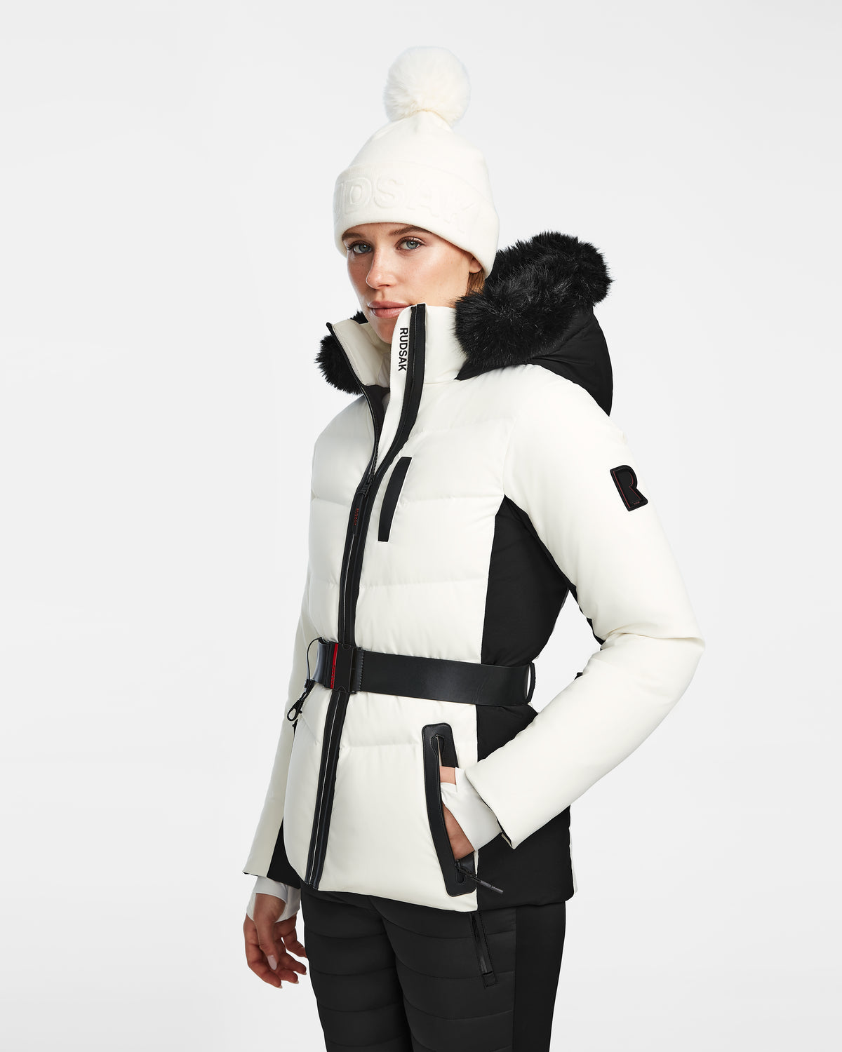 A woman models the Rudsak ALAIA - 8124816 Iceland white and black belted quilted ski jacket with a fur-trim hood, paired with black pants and a white knit pom-pom beanie, standing against a plain light background.