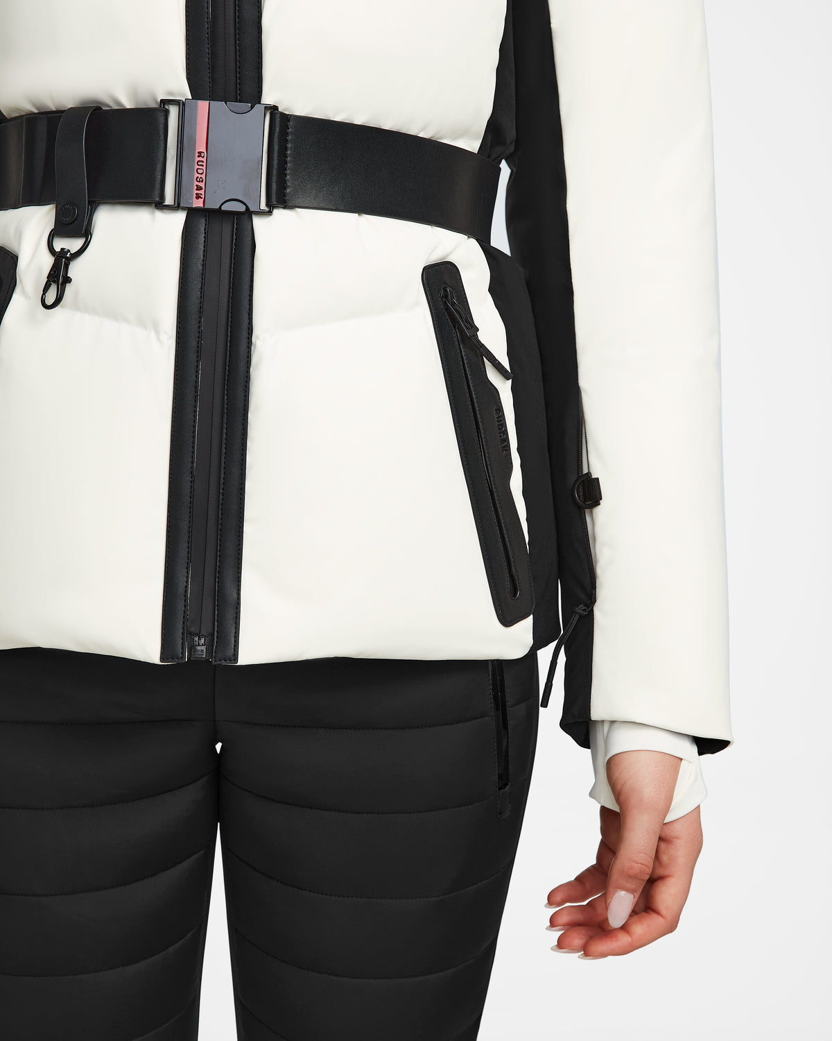 The model wears the Rudsak ALAIA - 8124816 Iceland ski jacket in white with black zippers and belt, paired with black quilted pants. Their right hand rests relaxed at their side.