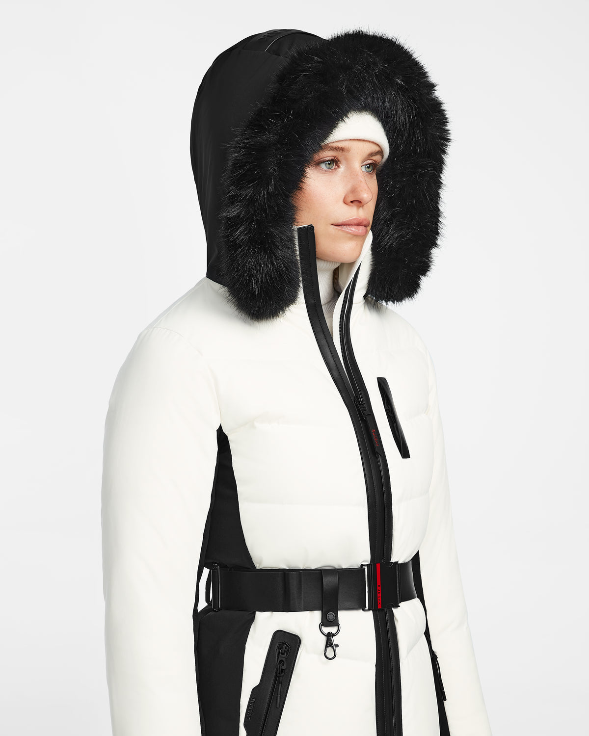 A person wearing the Rudsak ALAIA - 8124816 Iceland women’s quilted ski jacket with a fur-lined hood, paired with a white beanie and black belt, stands against a plain light background.