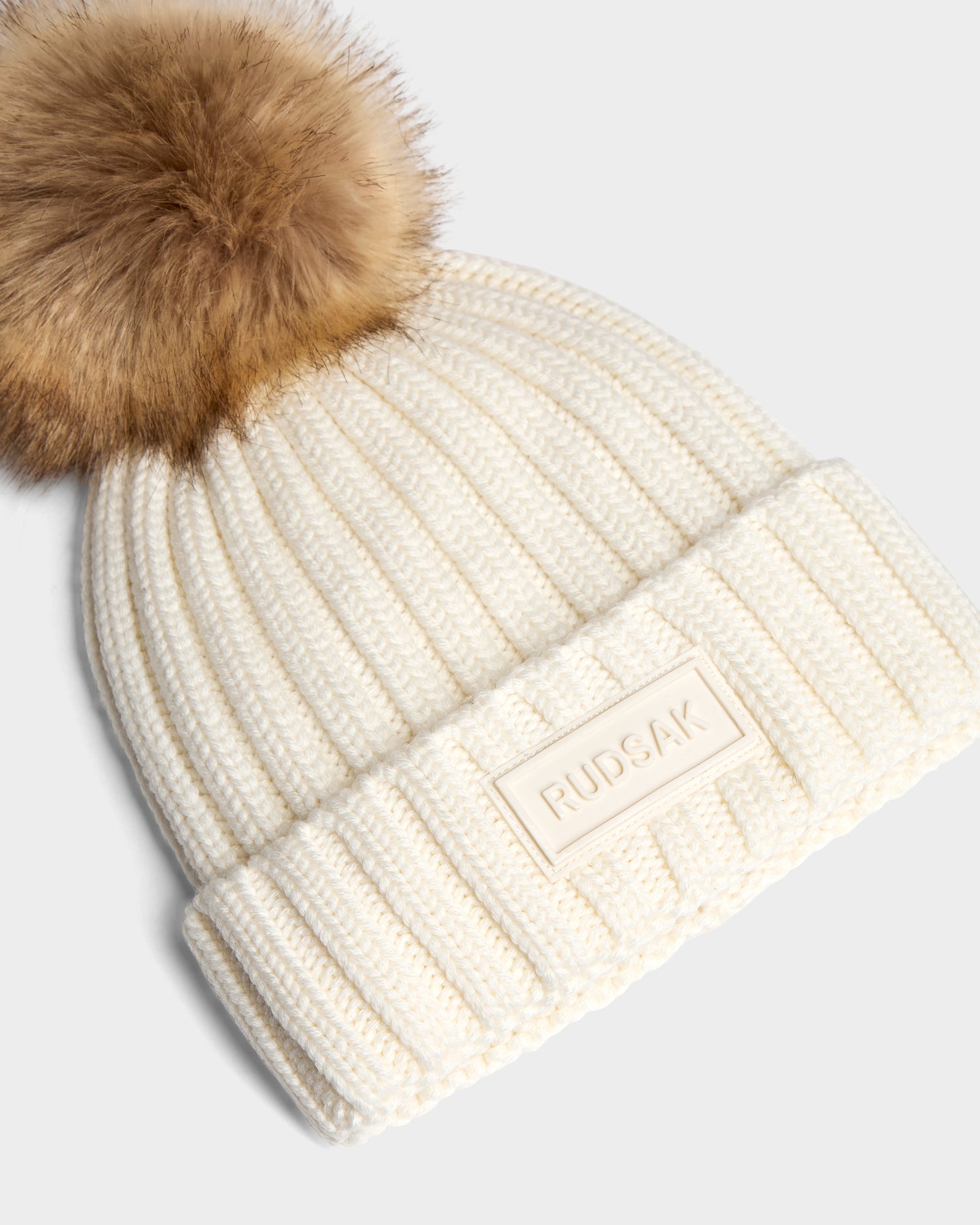 The ANISA - L423500 Optic White by Rudsak is a cream ribbed knit beanie with a folded cuff, large brown faux fur pom pom, and a rectangular patch featuring the Rudsak brand name—perfect for elevating your winter style.