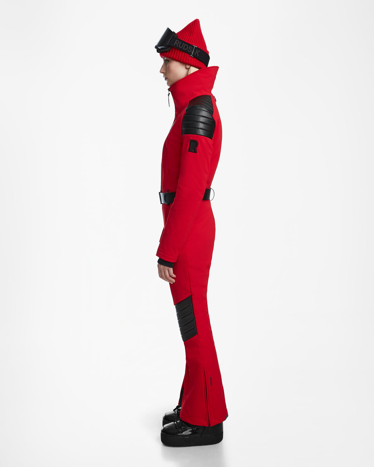 A person in profile wears the Rudsak ARLEY - 8124717 Flame Red fitted one-piece ski suit with black padded details, a matching red beanie, black ski goggles on the forehead, and black boots against a plain white background.