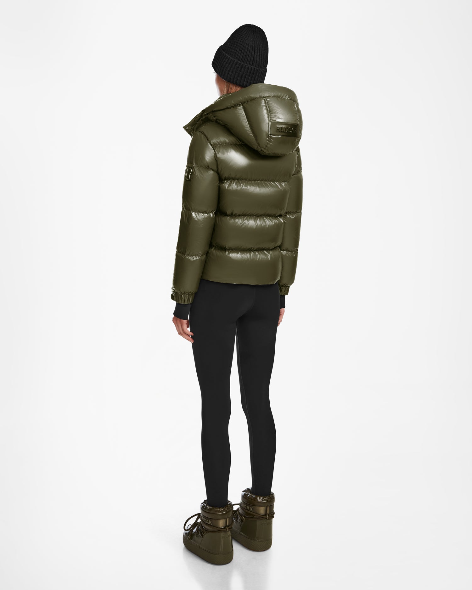 A person in the Rudsak ASHLYN S - 8124565 Loden Green puffer jacket, black leggings, chunky olive boots, and a black beanie stands facing away against a plain white background.