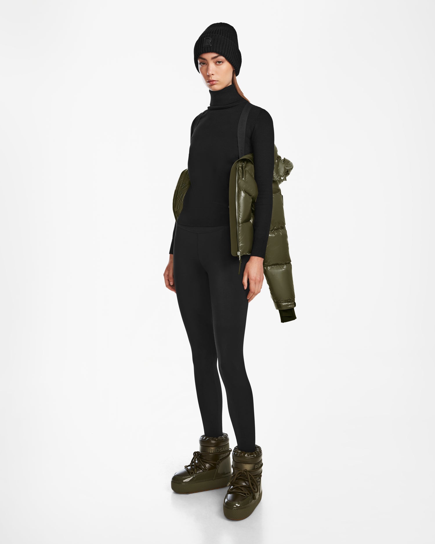 A person wearing Rudsak ASHLYN S - 8124565 Loden Green womens down puffer boots, a black beanie, black turtleneck, and leggings, with a water-resistant puffer jacket over one shoulder, stands against a plain white background.