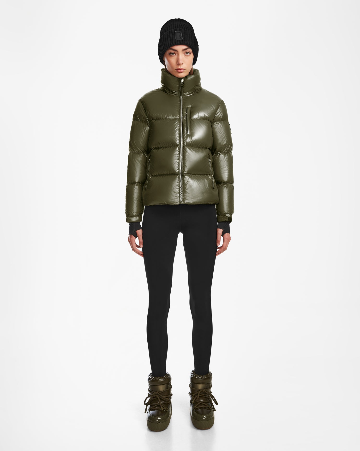 A person wearing the Rudsak ASHLYN S - 8124565 Loden Green puffer jacket stands against a plain white background, styled with black leggings, olive green winter boots, and a black knit beanie.