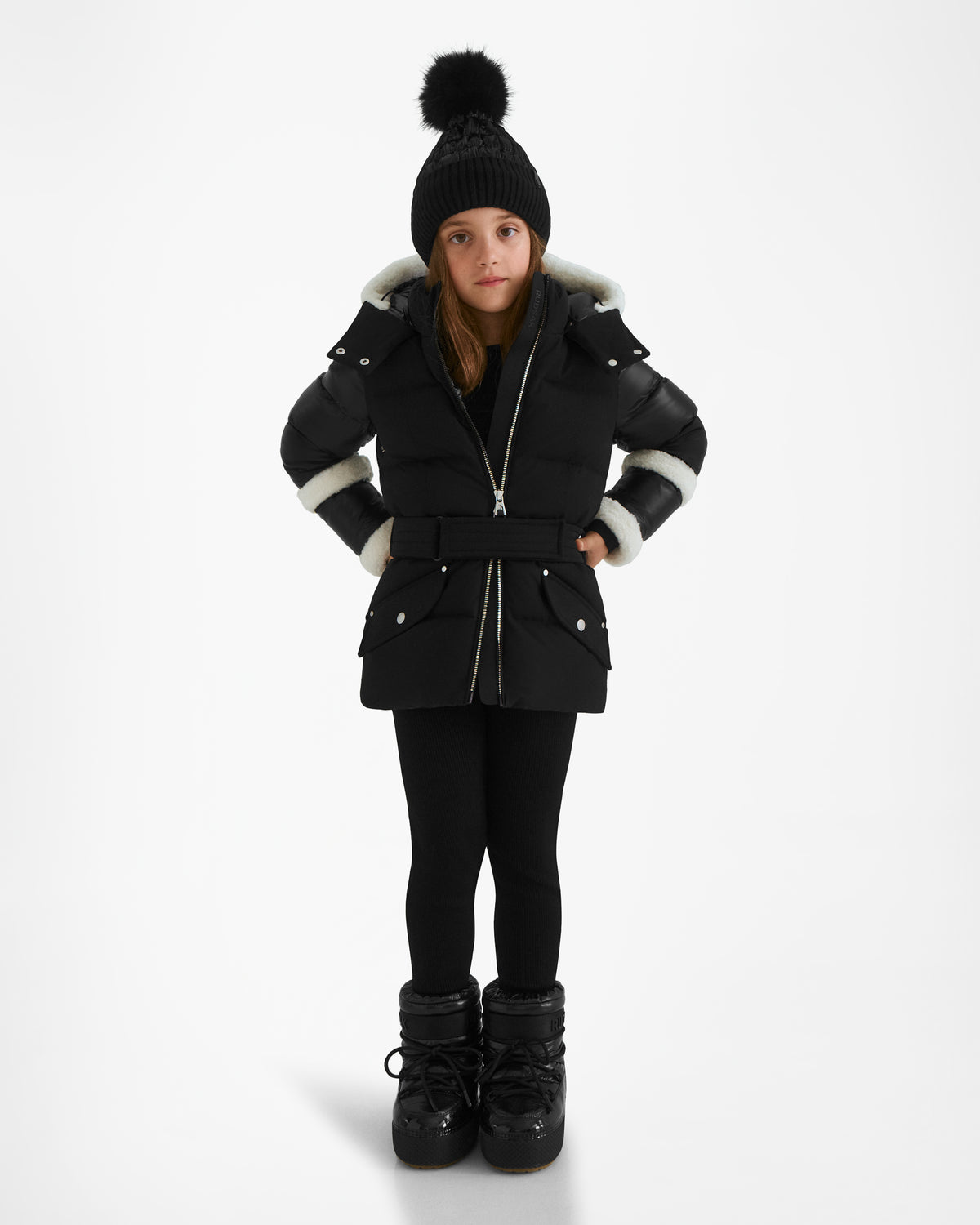 A young girl stands facing forward with her hands in her pockets, wearing the Rudsak AUDREY K - 0124504 Black down puffer jacket with a fur-trimmed, detachable hood, black leggings, boots, and a pom-pom knit hat.