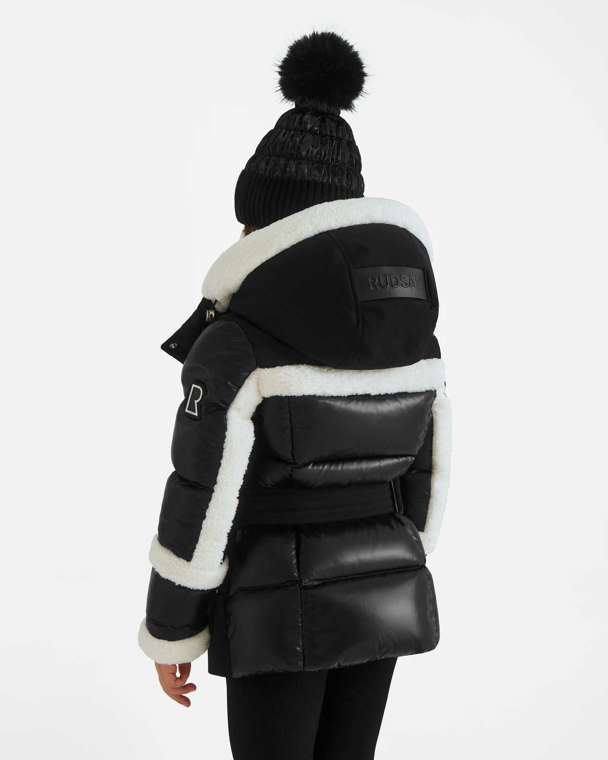 A person is seen from the back in a Rudsak AUDREY K - 0124504 Black girls down puffer jacket with white fleece trim and a water-repellent, detachable hood, paired with a black pom-pom beanie against a plain white background.
