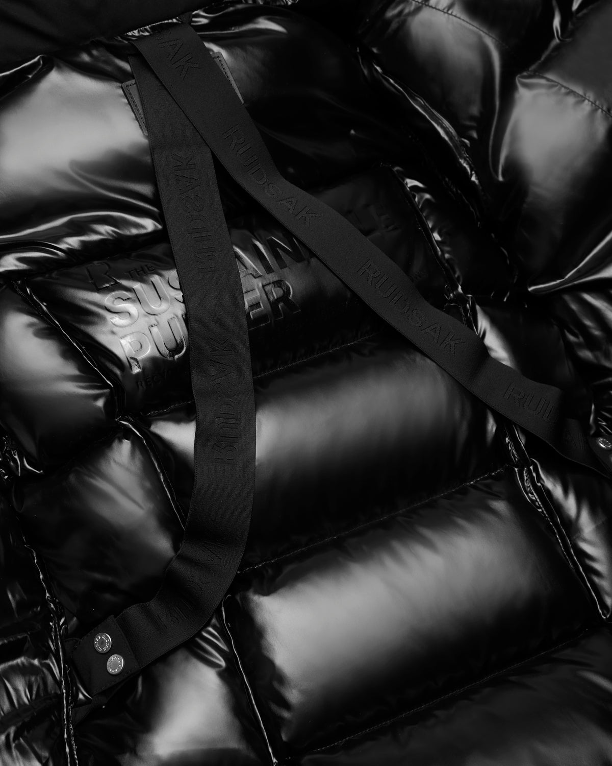Close-up of a shiny black AUDREY K - 0124504 puffer jacket by Rudsak, featuring water-repellent material, embossed text THE SUSTAIN RUNNER, and two matte black straps with RUINSAAK embossed.