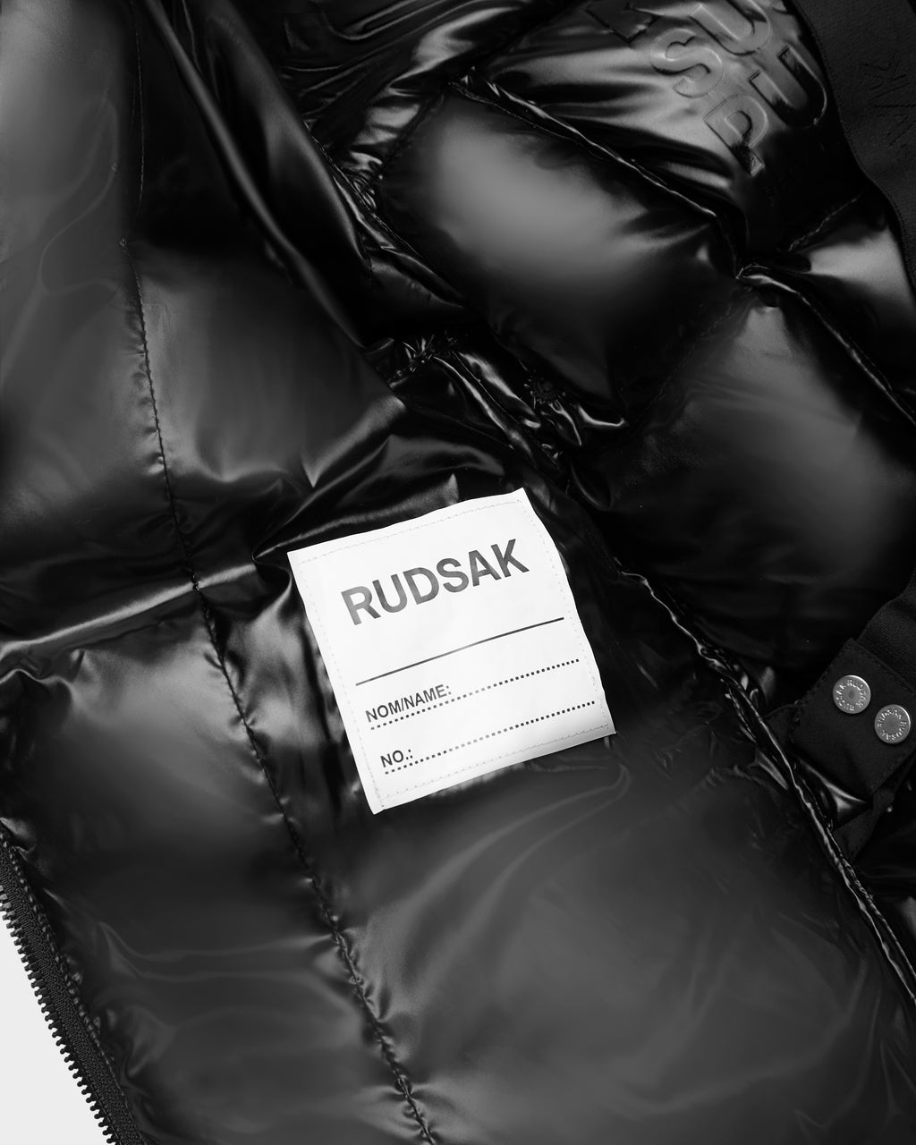 Close-up of the inside of a shiny black Rudsak AUDREY K - 0124504 girls down puffer jacket, featuring a white name and number label, stitched lining lettering, and water-repellent finish details.