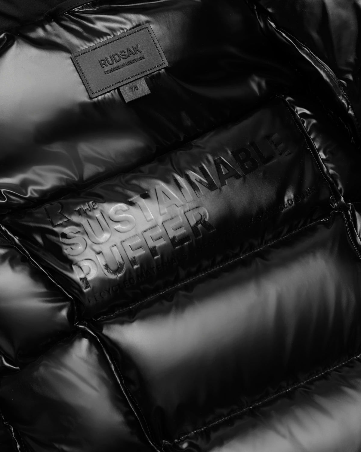 Close-up of Rudsaks AUDREY K - 0124504 black girls down puffer jacket featuring embossed THE SUSTAINABLE PUFFER text, a water-repellent finish, and a stitched Rudsak label at the top in size 7/8.