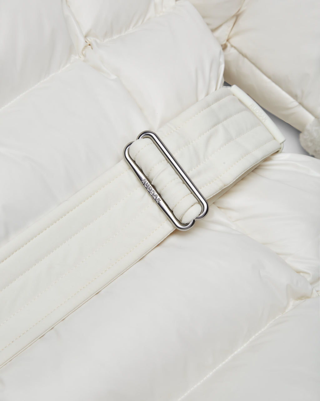 Close-up of the Rudsak AUDREY K - 0124504 Iceland white quilted belt with a silver rectangular buckle, displayed on matching water-repellent, padded fabric reminiscent of a wind-resistant puffer jacket.