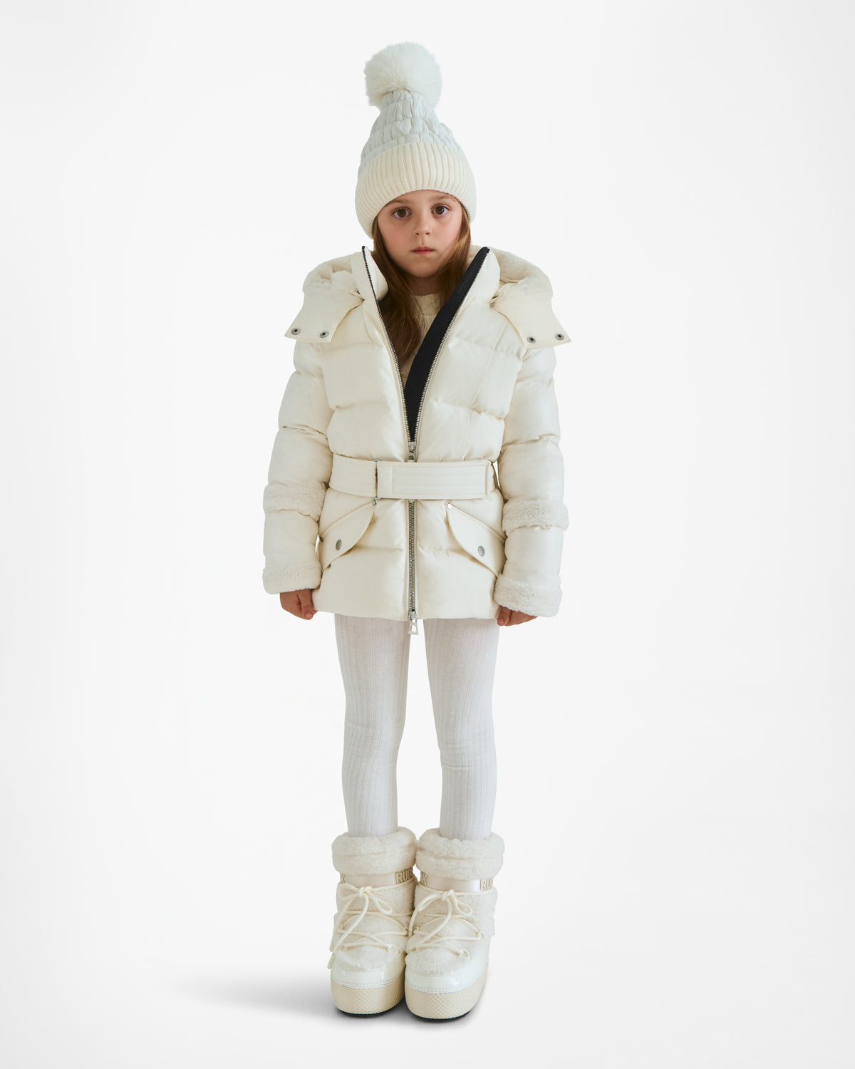 A young girl faces forward wearing the Rudsak AUDREY K - 0124504 Iceland girls down puffer jacket, paired with white leggings, winter boots, and a pom-pom beanie. She stands with a neutral expression against a plain white background.