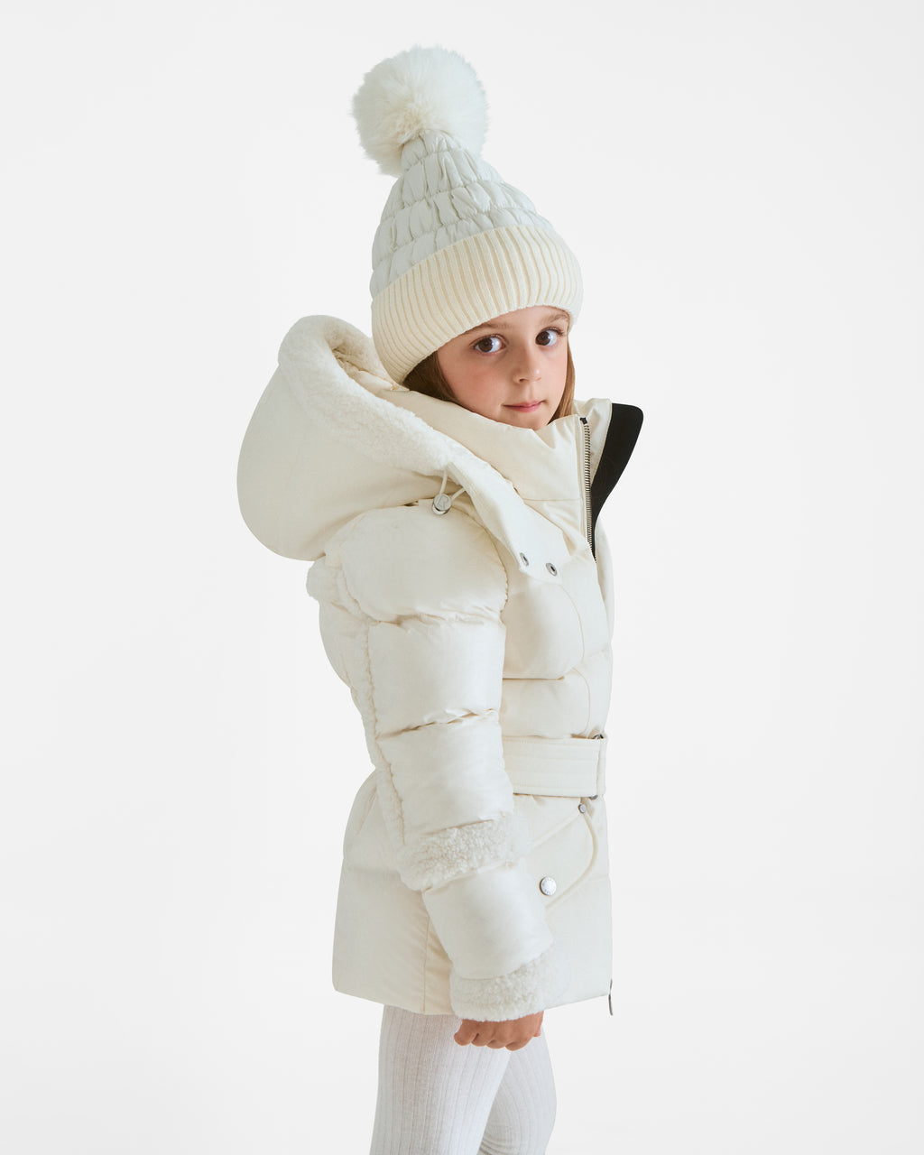 A young girl wears the Rudsak AUDREY K - 0124504 Iceland white down puffer jacket with a large hood and a matching pom-pom knit hat, standing against a plain white background. Water-repellent and wind-resistant for chilly days.