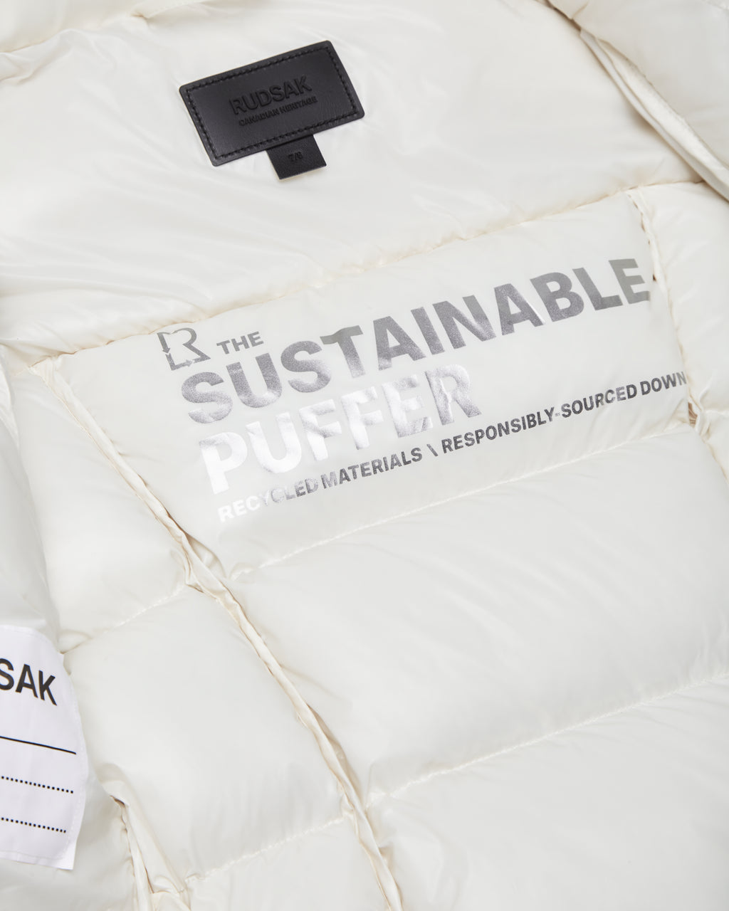 Close-up of the Rudsak AUDREY K - 0124504 Iceland girls white, wind-resistant down puffer jacket. THE SUSTAINABLE PUFFER—RECYCLED MATERIALS • RESPONSIBLY-SOURCED DOWN text and a black RUDSAK label are visible at the collar.
