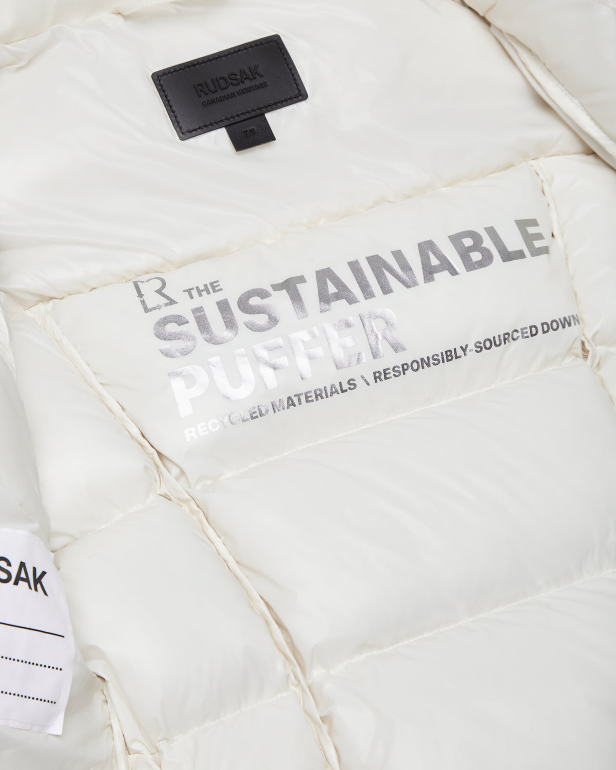 Close-up of the Rudsak AUDREY K - 0124504 Iceland girls white, wind-resistant down puffer jacket. THE SUSTAINABLE PUFFER—RECYCLED MATERIALS • RESPONSIBLY-SOURCED DOWN text and a black RUDSAK label are visible at the collar.