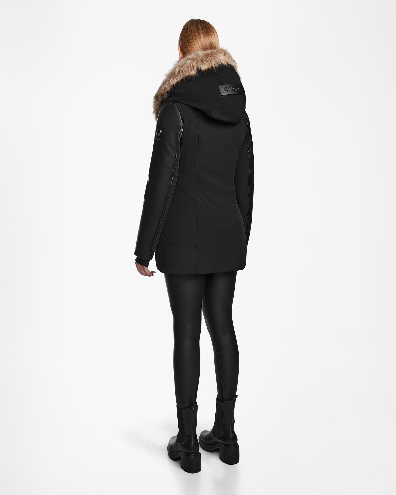 A person with long, light hair, seen from the back, wears Rudsak's AURORE R - L124535 Black weather-resistant jacket with faux fur hood, black leggings, and black boots against a plain white background.