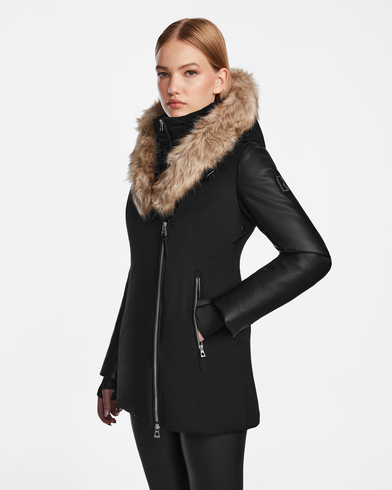 A woman with straight blonde hair models the Rudsak AURORE R - L124535 Black, a fitted winter coat with vegan leather sleeves and a tan faux fur-trimmed hood, standing against a plain white background and looking slightly to the side.