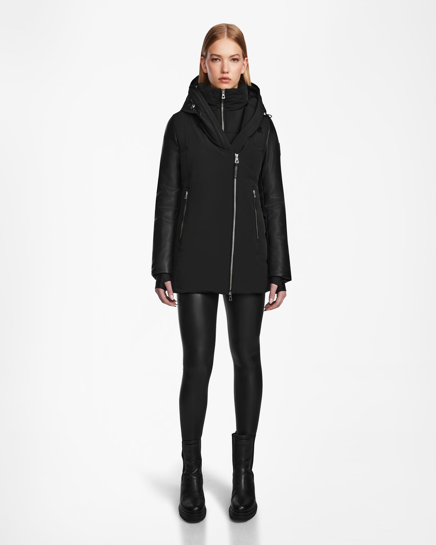 A woman faces forward against a white background, wearing the Rudsak AURORE R - L124535 Black weather-resistant jacket with vegan leather sleeves, paired with black leather-look leggings and black boots.