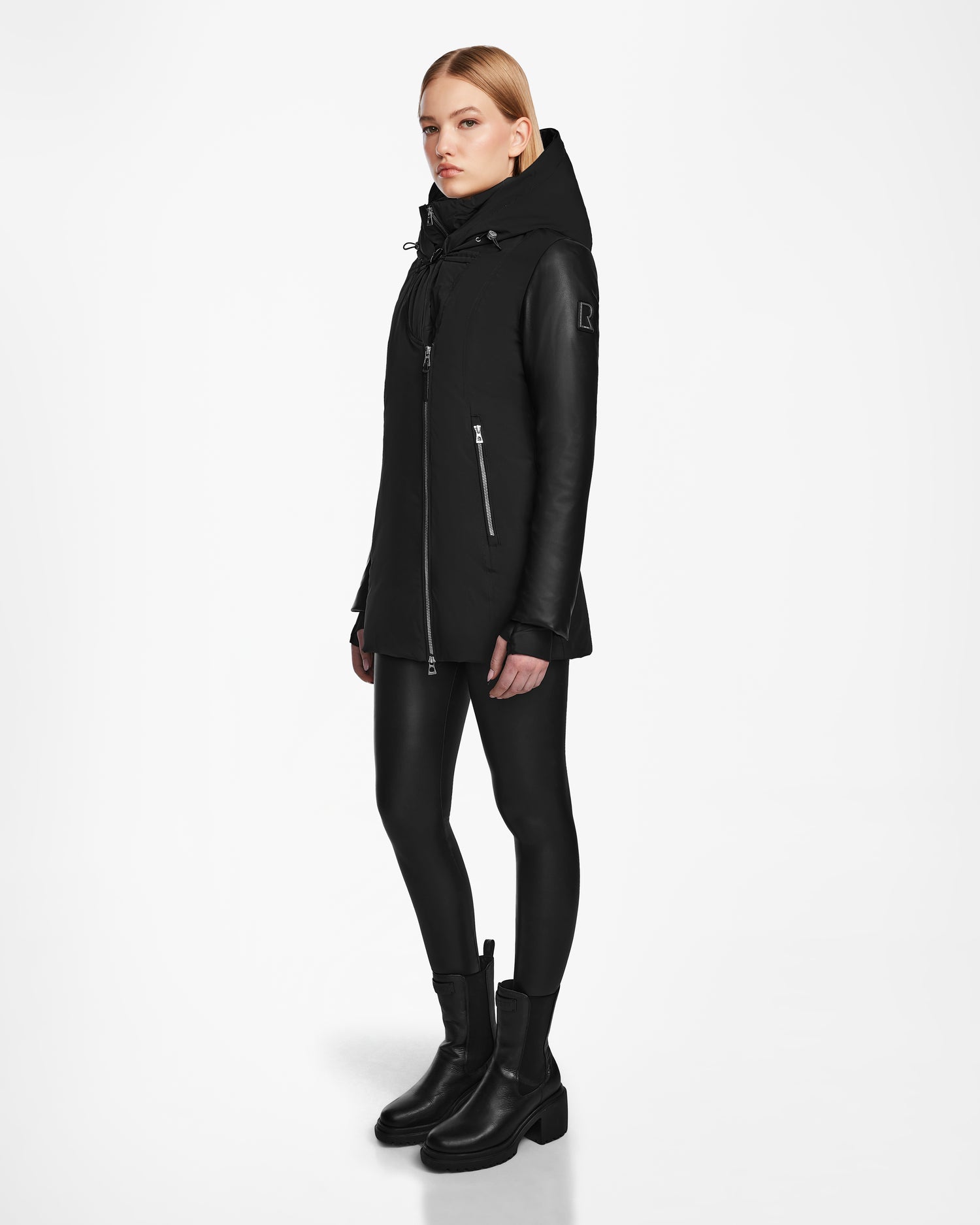 A woman stands against a white background wearing the Rudsak AURORE R - L124535 Black jacket with vegan leather sleeves, paired with black leggings and boots, looking slightly to the side with her arms relaxed.
