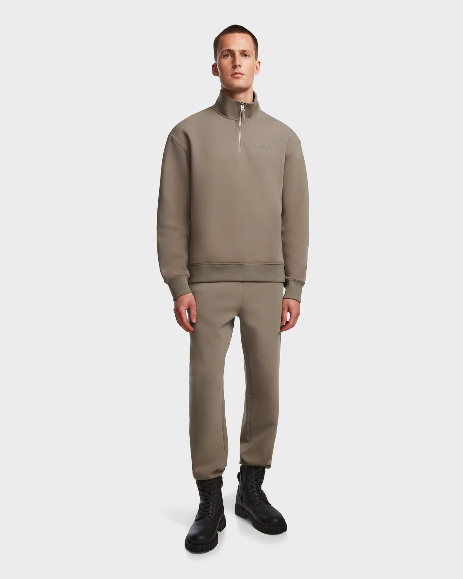 A person stands facing forward, wearing the Rudsak AVERY - 9524505 Cement M unisex mock neck zip-up sweater and matching jogger pants with black lace-up boots, against a plain white background.