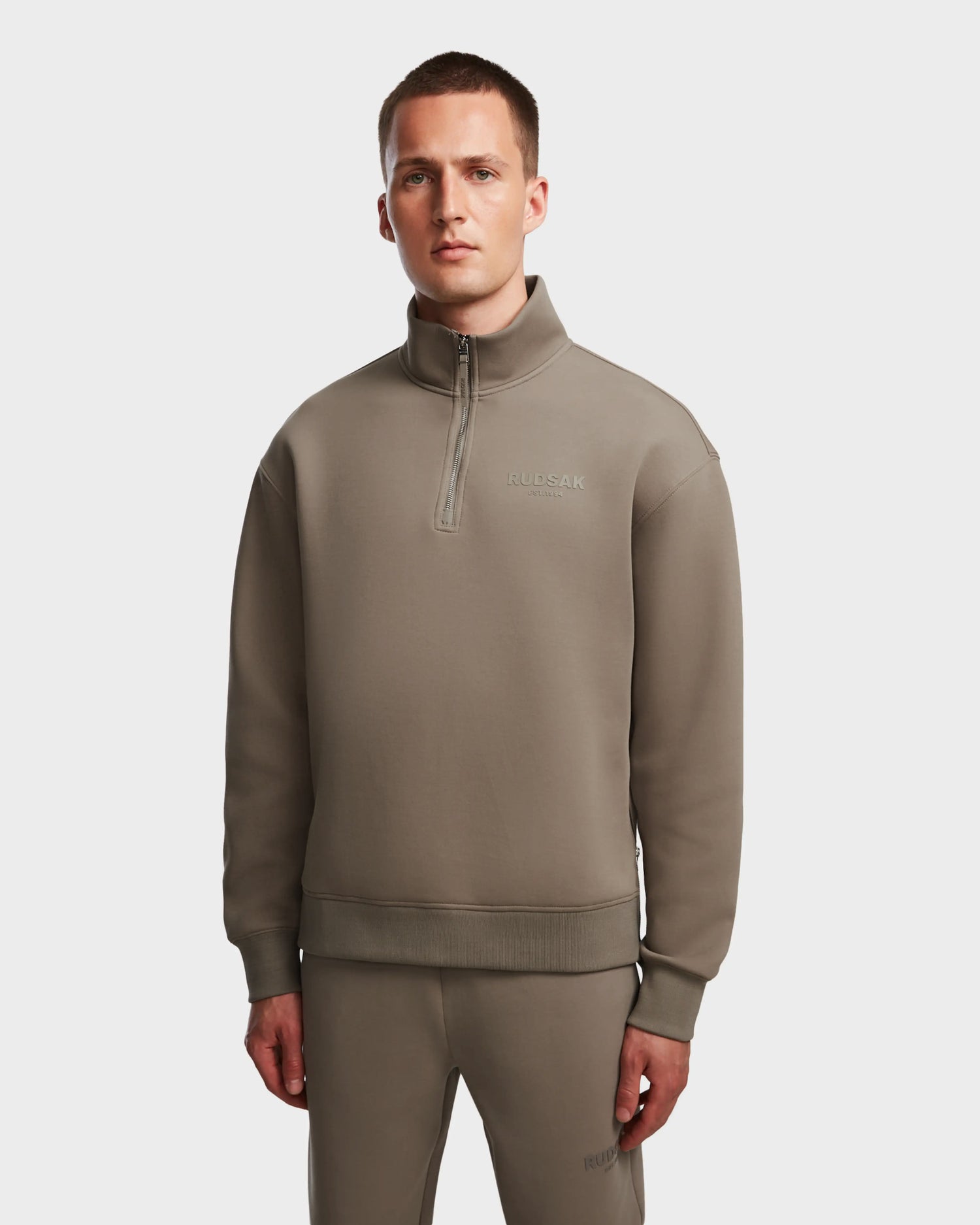 A man wearing the Rudsak AVERY - 9524505 Cement M taupe mock neck zip-up sweater and matching pants stands facing forward against a plain white background. The unisex sweater features RULSAAK subtly printed on the chest.