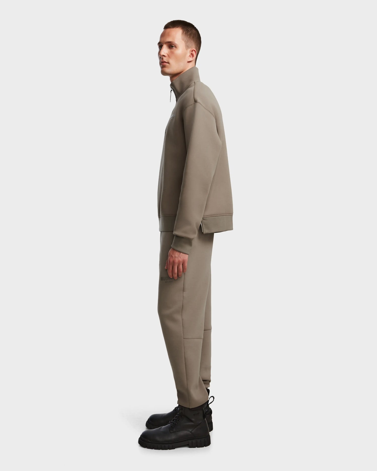 A man in profile wears the AVERY - 9524505 Cement M zip-up sweater by Rudsak with matching pants and black lace-up boots, standing against a plain light grey background.