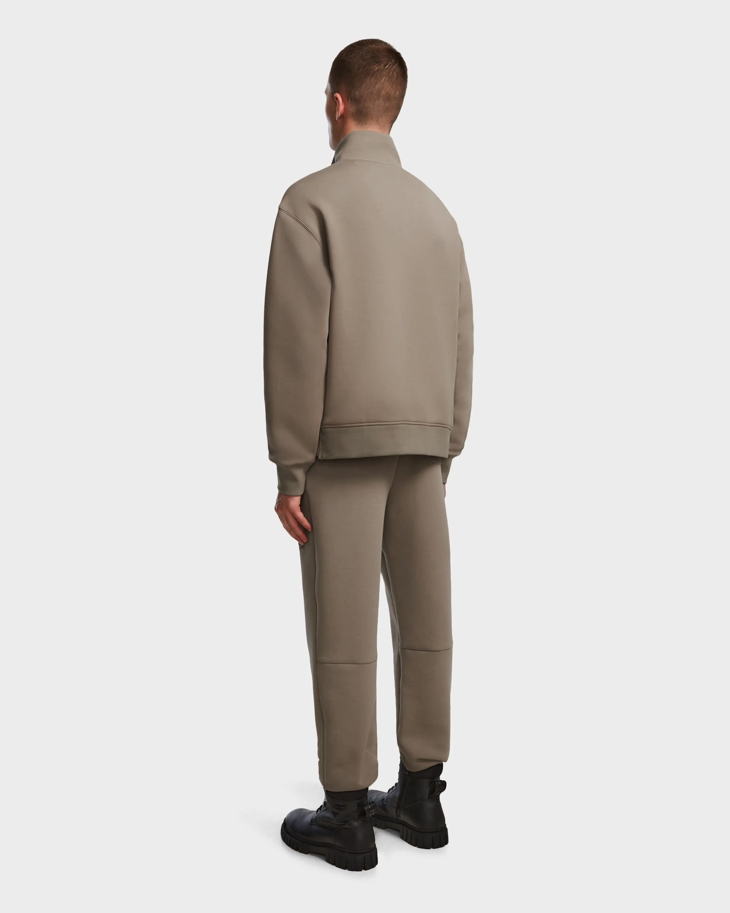 A person faces away, dressed in the Rudsak AVERY - 9524505 Cement M taupe mock neck zip-up sweater with matching pants and black chunky boots, standing against a plain white background.