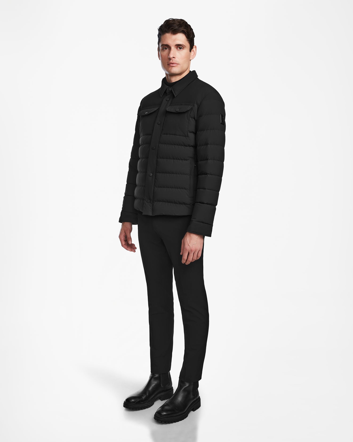 A man stands against a white background wearing the Rudsak AZIEL - M124521 Black mens puffer shirt jacket, styled with black pants, a black shirt, and black boots. He looks slightly to the side with a neutral expression.
