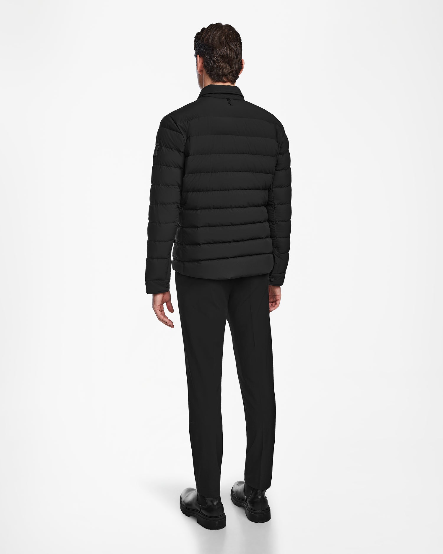 A man faces away, wearing the Rudsak AZIEL - M124521 Black mens puffer shirt jacket with duck down insulation, paired with black pants and shoes, set against a plain white background.