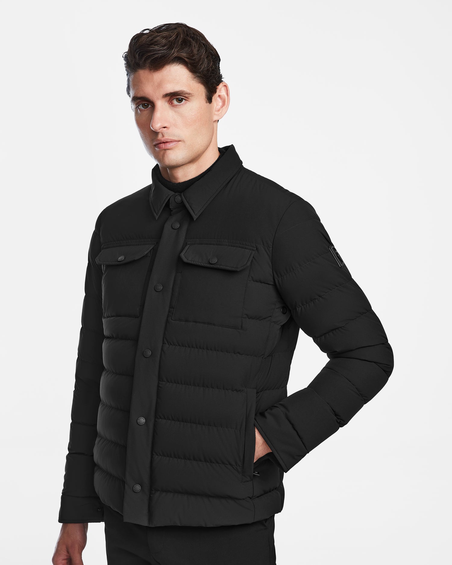 A man wearing the Rudsak AZIEL - M124521 Black puffer shirt jacket with front pockets stands against a white background, looking slightly to the side with one hand in his pocket.