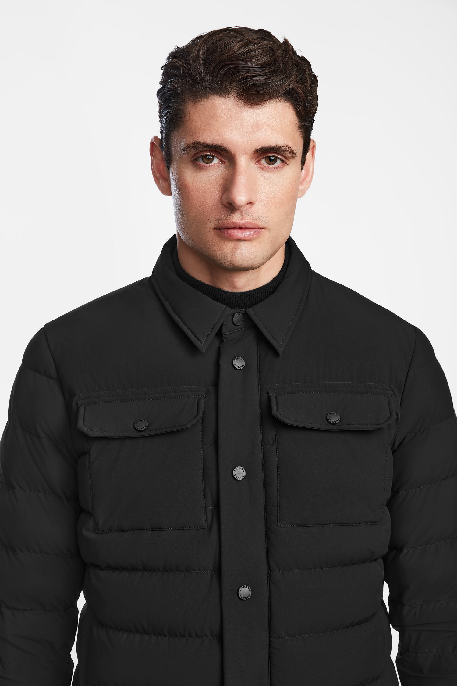 A man with short brown hair wears the Rudsak AZIEL - M124521 Black weather-resistant mens puffer shirt jacket with chest pockets, standing against a plain white background.