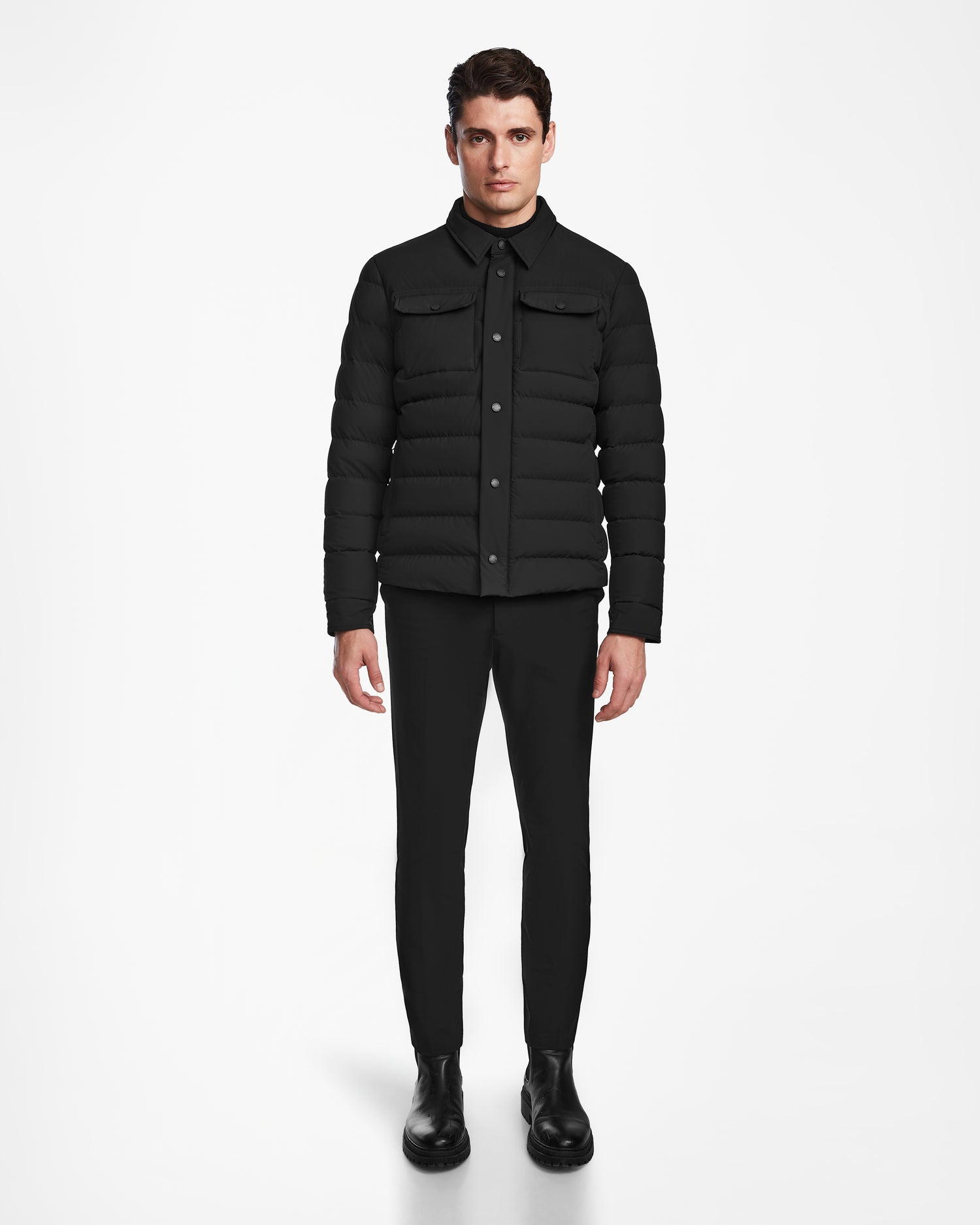 A man stands facing forward against a white background, wearing the Rudsak AZIEL - M124521 Black mens puffer shirt jacket, paired with black pants and boots. His arms are relaxed at his sides.