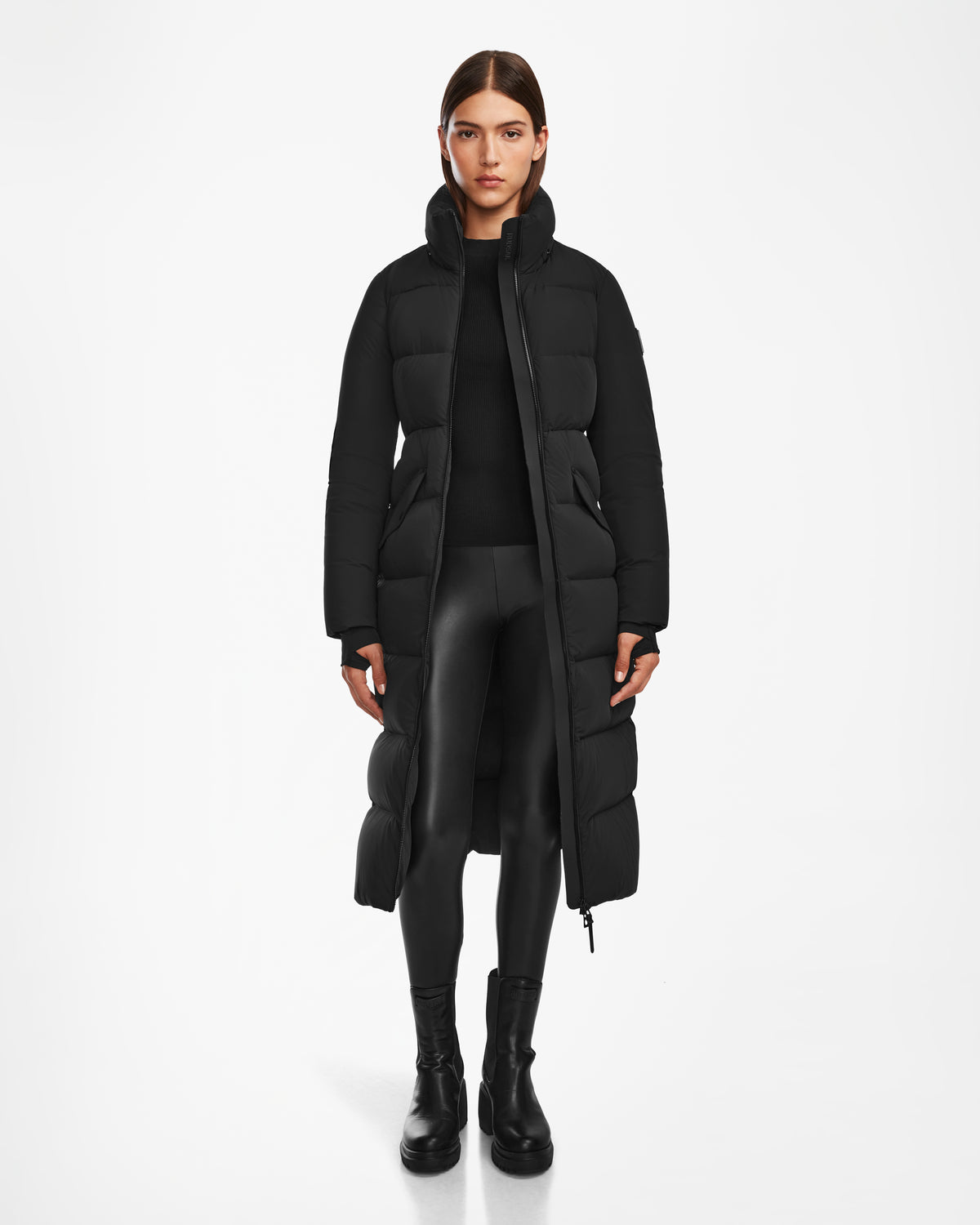A woman faces forward against a plain white background, wearing the Rudsak CALI - 8124600 Black womens long down puffer coat with black leggings and boots.
