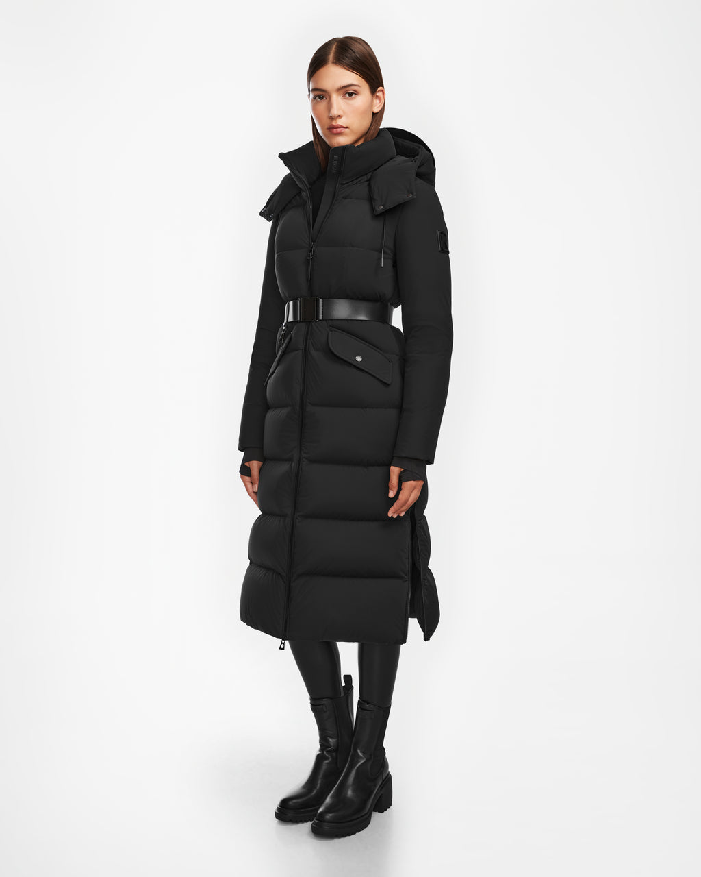 A woman wearing the Rudsak CALI - 8124600 Black long down puffer with belt, black leggings, and ankle boots stands against a white background; the jacket features a high collar and hood. She looks straight ahead with a neutral expression.